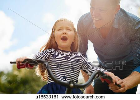 Stock Image - Father teaching daughter to ride bicycle. Fotosearch