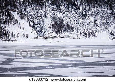 Stock Image - Frozen lake and village beneath snow covered mountain, Engadin, Switzerland. Fotosearch
