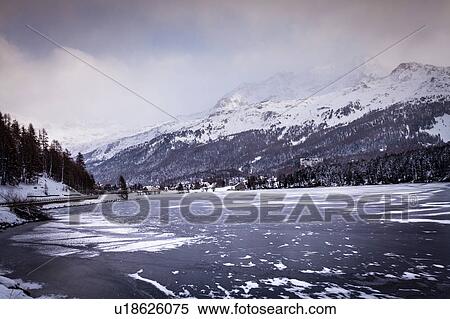 Stock Photography - Frozen lake and village beneath snow covered mountain, Engadin, Switzerland. Fotosearch
