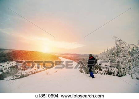 Picture - Man on snow covered landscape looking at view of sunset over frozen river. Fotosearch