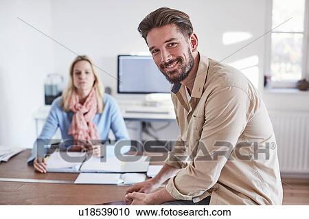 Stock Image - Man sitting on desk in office looking at camera smiling. Fotosearch