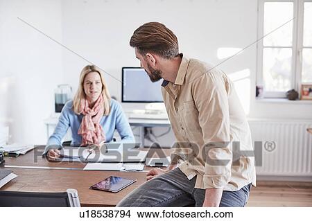 Man sitting on desk in office talking to colleague View Large Photo Image Picture - Man sitting on desk in office talking to colleague. Fotosearch