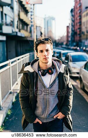 Stock Image - Man with headphones in urban area, hands in pockets looking at camera. Fotosearch
