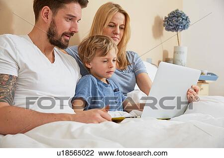 Stock Image - Mother and father in bed with son using digital tablet. Fotosearch
