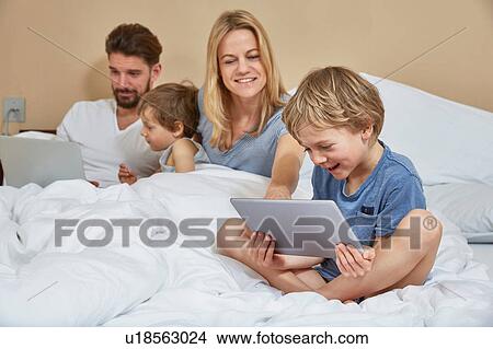 Mother and father in bed with sons using technology View Large Photo Image Picture - Mother and father in bed with sons using technology. Fotosearch