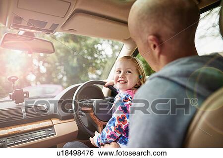 Stock Photograph - Over shoulder view of daughter sitting on father lap driving car. Fotosearch