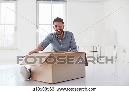Stock Image - Portrait of young man sitting on floor opening cardboard box in new house. Fotosearch