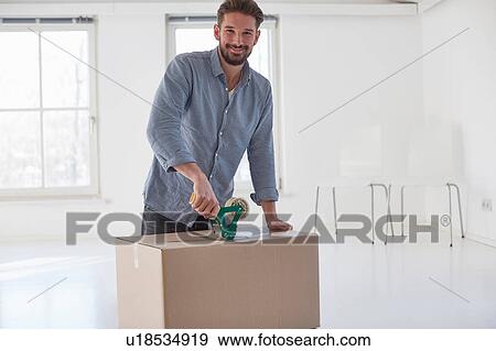 Stock Photo - Portrait of young man taping cardboard box whilst moving house. Fotosearch