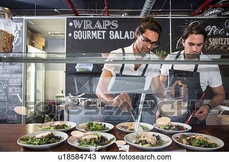 Stock Image - Restaurateurs preparing salad behind service counter. Fotosearch