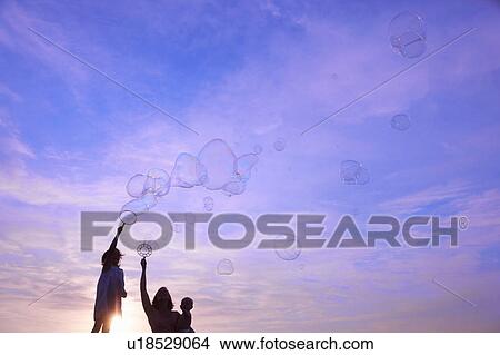 Silhouetted woman with baby son and daughter blowing bubbles against blue sky, Buonconvento, Tuscany, Italy View Large Photo Image Picture - Silhouetted woman with baby son and daughter blowing bubbles against blue sky, Buonconvento, Tuscany, Italy. Fotosearch