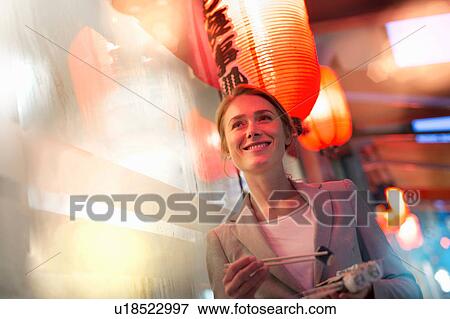 Stock Photo - Woman eating takeaway sushi whilst strolling on city street at night. Fotosearch