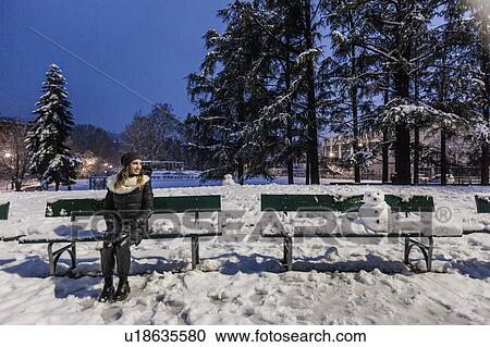 Stock Image - Woman sitting on park bench in covered park in the evening. Fotosearch