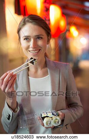 Stock Image - Woman strolling and eating takeaway sushi on city street at night. Fotosearch