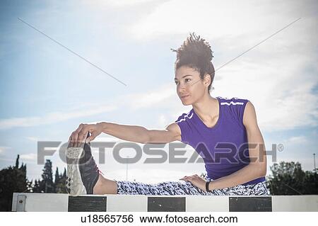 Stock Photograph - Young female hurdler touching toes on hurdle at sport facility. Fotosearch