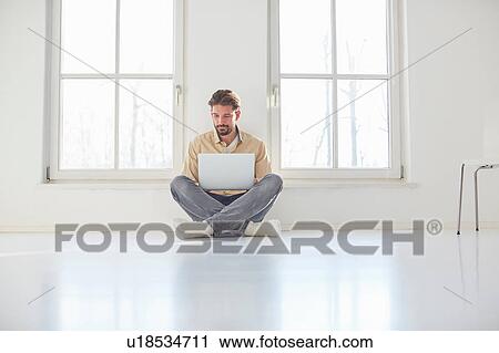 Stock Image - Young man sitting on floor browsing laptop in empty new home. Fotosearch