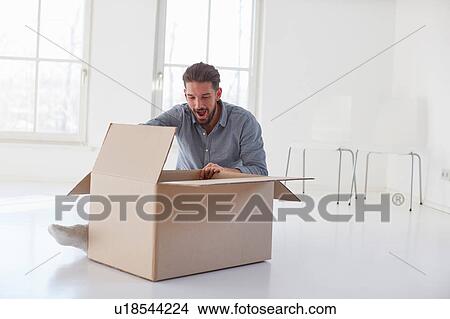 Picture - Young man sitting on floor opening cardboard box in new house. Fotosearch