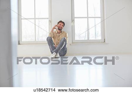 Picture - Young man sitting on floor talking on smartphone in empty new home. Fotosearch