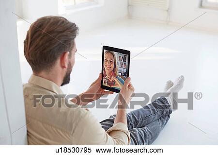 Stock Photography - Young man sitting on floor talking to girlfriend on digital tablet video call. Fotosearch