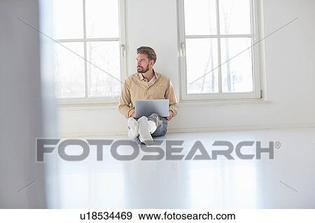 Stock Photo - Young man sitting on floor with laptop in empty new home. Fotosearch