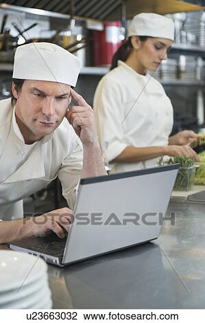 Stock Image - Chef using laptop and cook preparing food in commercial kitchen. Fotosearch