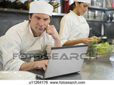 Stock Photograph - Chef using laptop and cook preparing food in commercial kitchen. Fotosearch
