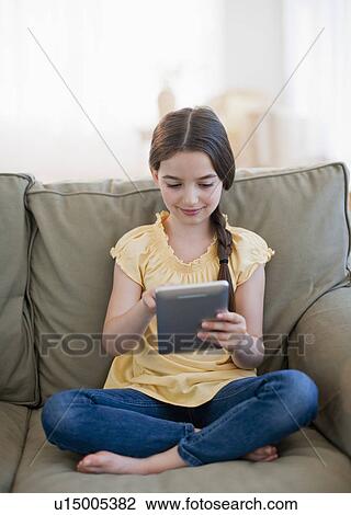 Stock Image - Girl (8-9) sitting on sofa and using digital tablet. Fotosearch