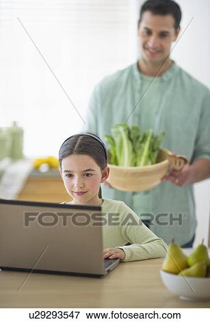 Stock Photo - Girl (8-9) using laptop and father preparing food in domestic kitchen. Fotosearch