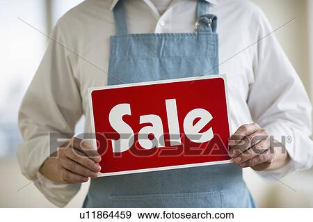 Stock Photo - Man wearing apron holding "sale" sign, mid section. Fotosearch