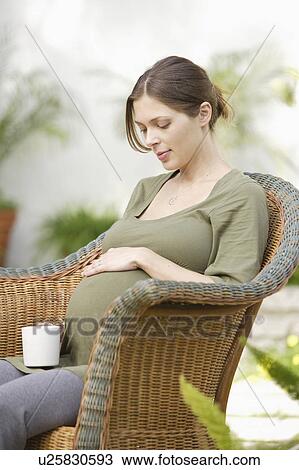 Stock Image - Pregnant woman sitting in wicker chair, touching belly. Fotosearch