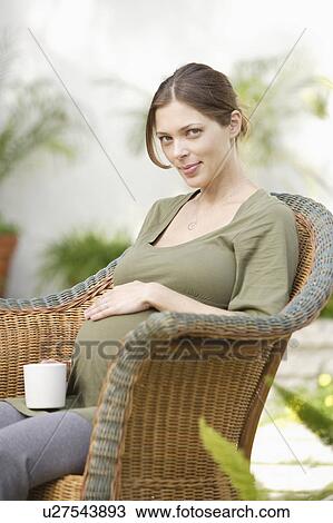 Stock Image - Pregnant woman sitting in wicker chair, touching belly. Fotosearch