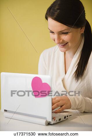 Stock Image - Smiling young woman using laptop with pink heart. Fotosearch