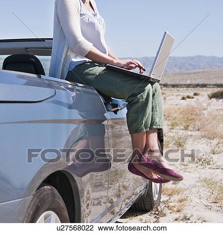 Stock Image - USA, California, Palm Springs, Woman sitting on side of convertible car in desert, using laptop. Fotosearch