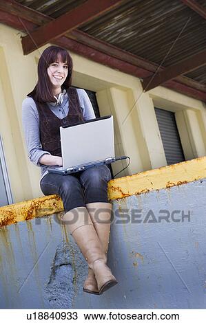 Stock Image - USA, New Jersey, Jersey City, young woman sitting on wall using laptop. Fotosearch