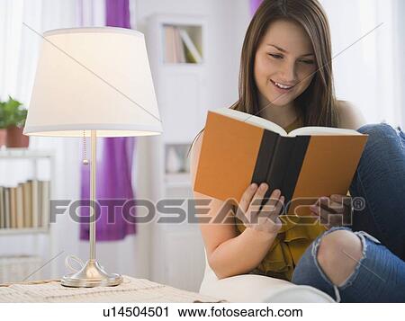 Stock Image - Young woman reading book in living room. Fotosearch