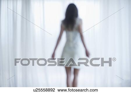 Stock Image - Young woman standing and looking through window. Fotosearch