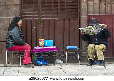 Stock Photo - Man reading a newspaper at a market stall with a woman sitting beside him, Sacred Valley, Cusco Region, Peru. Fotosearch