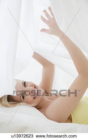 Stock Image - Blonde woman hiding under bed sheet. Fotosearch