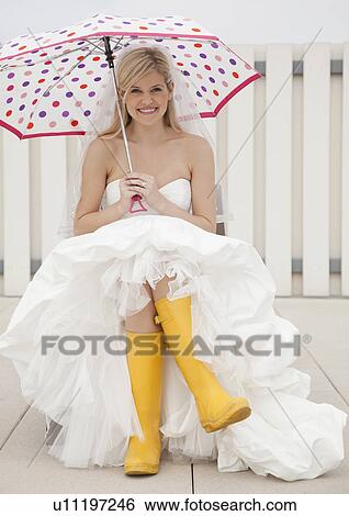 Stock Photograph - Bride sitting in yellow rubber boots with umbrella. Fotosearch
