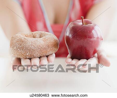 Stock Image - Close up of woman's hands holding donut and apple. Fotosearch