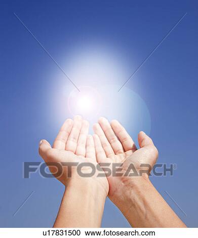 Stock Image - Close up of woman's hands holding sunlight on blue sky. Fotosearch