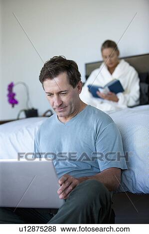 Stock Photo - Mature man with laptop in bedroom and woman reading book in bed. Fotosearch