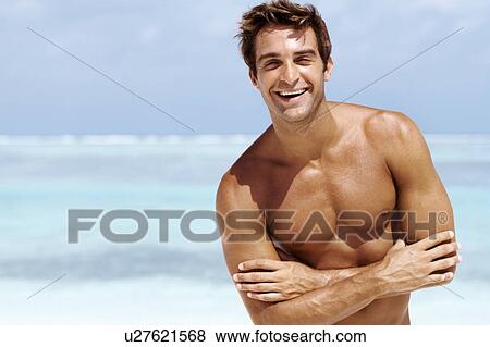 Stock Photo - Portrait of happy shirtless young male standing with folded hands at beach. Fotosearch