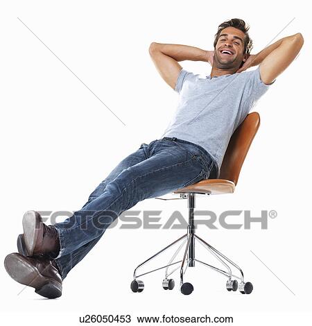 Stock Image - Portrait of young man relaxing on chair with hands behind head and smiling against white background. Fotosearch