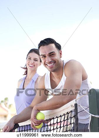 Stock Image - Smiling couple standing near tennis net. Fotosearch