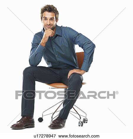 Stock Photo - Studio shot of young business man sitting on chair with hand on chin and smiling. Fotosearch