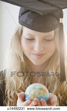 Stock Image - Woman wearing graduation gown holding small globe. Fotosearch