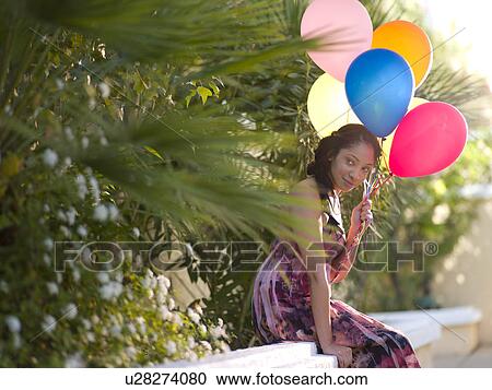 Stock Image - Young smiling woman sitting on wall and holding balloons. Fotosearch