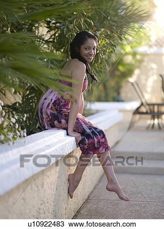 Stock Image - Young smiling woman sitting on wall. Fotosearch