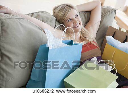 Stock Image - Young woman sitting on sofa surrounded by shopping bags. Fotosearch