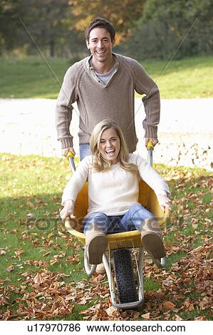 Stock Photograph - Man pushing wife through autumn leaves on wheelbarrow. Fotosearch